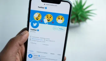 Una mano con un smartphone muestra el perfil oficial de Twitter con una foto de portada de emojis enmascarados, evocando recuerdos de los tiempos de 140 caracteres. Se ve una planta borrosa al fondo.