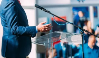 Una persona con un traje azul se encuentra de pie frente a un podio transparente con un micrófono, gesticulando con una mano mientras se dirige a una audiencia sobre Acción en la Política.