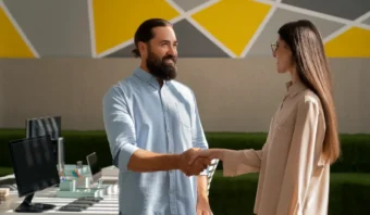 Un hombre y una mujer están de pie en una oficina, sonriendo y dándose la mano, lo que refleja una experiencia de cliente exitosa. Al fondo se ven monitores de computadora y escritorios.