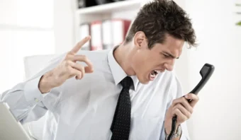 Un hombre con camisa blanca y corbata está sentado en un escritorio, gritando enojado en un teléfono fijo mientras levanta un dedo, como si dijera: "El cliente no importa".