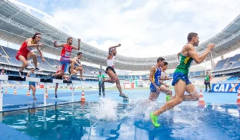 Varios atletas masculinos compiten en una carrera de obstáculos, saltando una barrera de agua en una pista azul dentro de un gran estadio durante los Juegos Olímpicos sin Televisa.