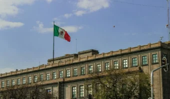 Una gran bandera mexicana ondea sobre un histórico edificio de piedra bajo un cielo parcialmente nublado, evocando la última estocada y la agonizante imagen presidencial. Árboles y un semáforo se alzan en primer plano.