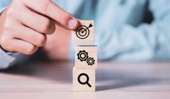 Una mano coloca un bloque de madera con un ícono de objetivo Objetivo SMART encima de dos bloques que muestran íconos de engranaje y lupa, apilados sobre una mesa.