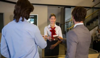 Una mujer detrás de un mostrador, vestida con una camisa blanca y una bufanda roja, sonríe y se muestra amable con mi cliente mientras entrega documentos a dos hombres que están parados al otro lado.