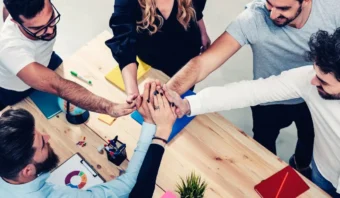 Cinco personas de pie alrededor de una mesa se toman de las manos en el centro, mostrando el trabajo en equipo y el poder de la participación en un entorno de oficina con computadoras portátiles, papeles y material de oficina visibles.