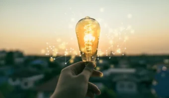 Una mano sostiene una bombilla encendida al aire libre al atardecer, con lÃ­neas de red digital superpuestas alrededor de la bombilla, simbolizando orientaciÃ³n a resultados y pensamiento innovador.