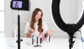 Una mujer se sienta en una mesa con productos de maquillaje, gesticulando con las manos mientras es filmada por una cÃ¡mara e iluminada por un anillo de luz, una escena comÃºn entre los influencers que crean una guÃ­a prÃ¡ctica para entusiastas de la belleza.