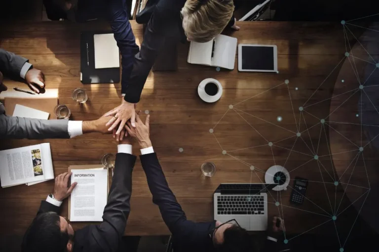 Cinco personas sentadas alrededor de una mesa de madera juntan sus manos en el centro, celebrando un networking exitoso; sobre la mesa son visibles papeles, una computadora portátil, café y una tableta.