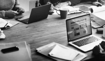 Personas sentadas alrededor de una mesa de madera con computadoras portátiles, documentos, tazas de café y material de oficina, participando en una reunión. La pantalla de una computadora portátil muestra una estrategia de marketing según la línea del marketing de experiencia.