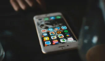 Un teléfono inteligente que muestra varios íconos de aplicaciones coloridas reposa sobre una superficie oscura, evocando una atmósfera de Black Mirror, con una mano borrosa y un vidrio visibles cerca.