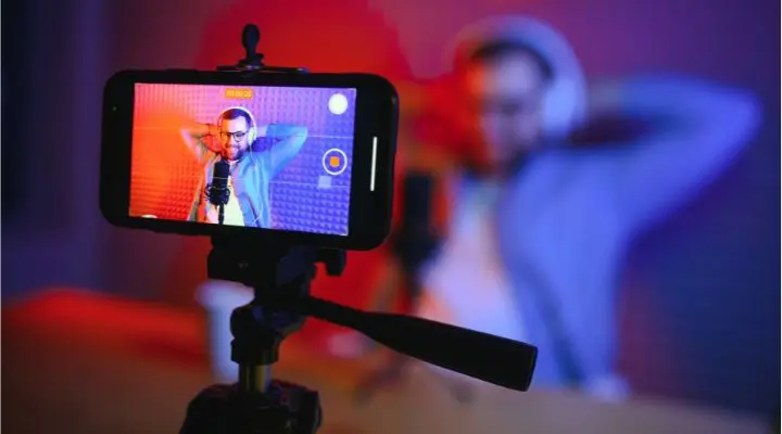 Una persona con auriculares se sienta en un escritorio con un micrófono y se graba con un teléfono inteligente para una sesión en vivo de Facebook live en un estudio colorido.