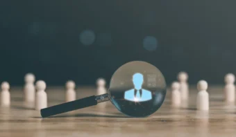 Una lupa se enfoca en la silueta digital de una persona con traje entre figuras de madera sobre una mesa, simbolizando la selección o análisis personal en el proceso de reclutamiento.