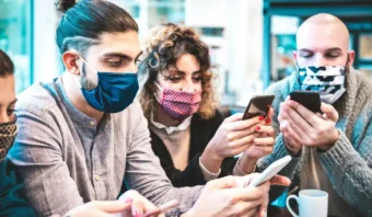 Cuatro nativos digitales con mascarillas se sientan juntos en el interior, mirando atentamente sus teléfonos inteligentes.