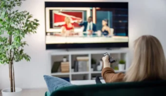 Una persona sentada en un sofá sostiene un control remoto y mira Buenos Comerciales durante un programa de noticias en un televisor de pantalla plana en una sala de estar con una planta en maceta.