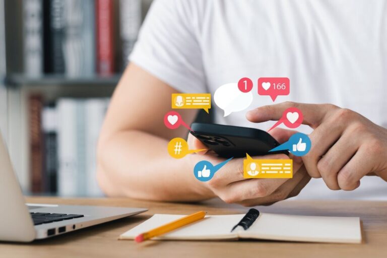 Una persona sostiene un smartphone con varios iconos de notificaciones de redes sociales (me gusta, comentarios y seguidores) flotando sobre el dispositivo, lo que ilustra cómo elegir influencers. Un lápiz y un cuaderno descansan sobre la mesa.