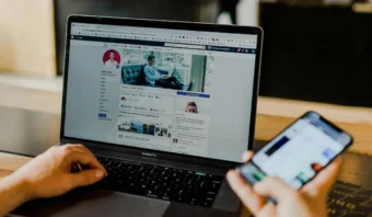 Persona que usa una computadora portátil para ver un perfil de Facebook mientras sostiene un teléfono inteligente en su escritorio, posiblemente preparándose para ver Facebook Live.