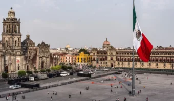 Una gran bandera mexicana se encuentra en el centro de la plaza del Zócalo en la Ciudad de México, donde el presidente a menudo se dirige a la nación, rodeado de edificios históricos y gente caminando.