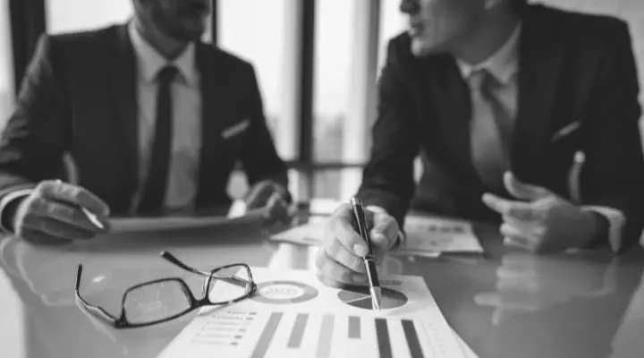 Dos hombres de traje se sientan en una mesa durante la entrevista, revisando cuadros y gráficos; uno señala un documento con un bolígrafo, mientras un par de gafas descansa sobre la mesa.
