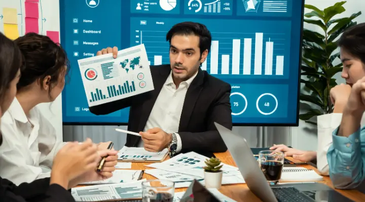 Un hombre de traje, un mercadólogo, presenta gráficos de datos a tres colegas en una sala de reuniones, con gráficos mostrados en una pantalla grande detrás de ellos.