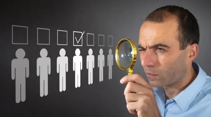 Un hombre sostiene una lupa y mira Ã­conos de personas, uno de ellos marcado con una marca de verificaciÃ³n, que simboliza el proceso para elegir agencia.