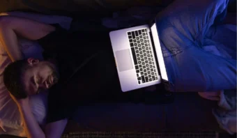 Un hombre yace en un sofá con el brazo bajo la cabeza, durmiendo, mientras una computadora portátil abierta descansa sobre su regazo, con la pantalla brillante; tal vez se quedó dormido mientras transmitía algo relajante en la plataforma Napflix.
