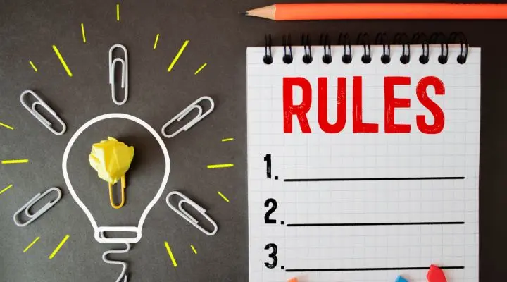 Un bloc de notas con la palabra "REGLAS" y una lista numerada se encuentra junto a un lápiz y un contorno de bombilla hecho con clips sobre una superficie oscura, simbolizando ideas para Las reglas.