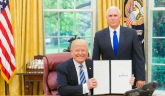 Un hombre sentado en un escritorio sostiene un documento firmado mientras Donald Trump está junto a él en una oficina con banderas y una gran ventana.