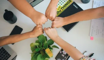 Cuatro personas demuestran trabajar en equipo con un choque de puños grupal sobre un escritorio blanco, rodeado de cuadernos, tazas de café, una computadora portátil y una planta en maceta.