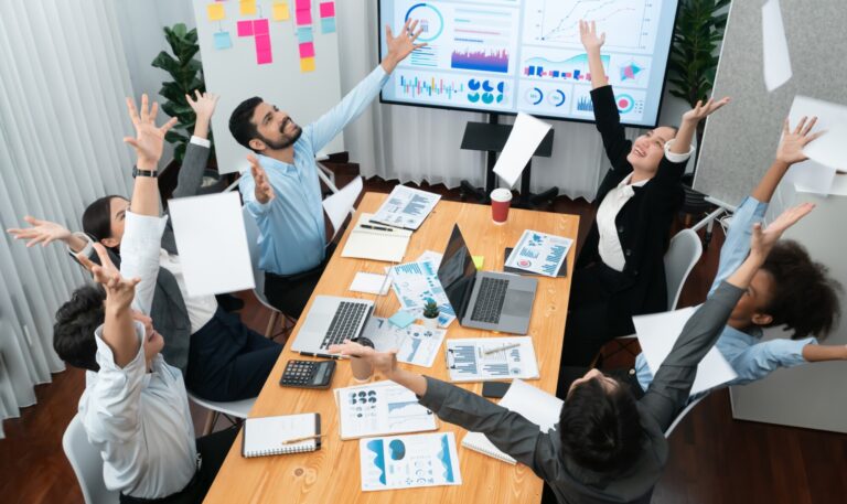 Seis personas vestidas de negocios se sientan alrededor de una mesa de conferencias, lanzando papeles al aire, celebrando el éxito de trabajar en equipo, con computadoras portátiles, cuadros y gráficos visibles sobre la mesa y en el fondo.