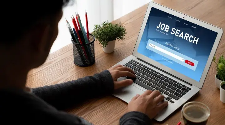 Una persona usa una computadora portátil que muestra un sitio web de búsqueda de empleo para encontrar trabajo en un escritorio con una taza de café, plantas en macetas y lápices.
