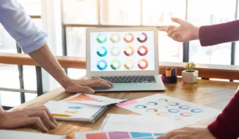 Dos personas discuten sobre gráficos de anillos coloridos exhibidos en una computadora portátil, con gráficos impresos y cuadernos en un escritorio de madera en un entorno de oficina iluminado, perfecto para intercambiar ideas para crear una marca.