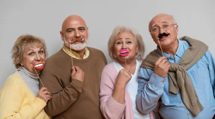 Cuatro miembros de la generaciÃ³n del baby boom estÃ¡n juntos, sonriendo y sosteniendo divertidos adornos de papel con forma de labios y bigotes frente a sus caras sobre un fondo liso Early Boomers.