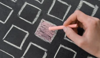 Una mano usa tiza rosa para colorear un cuadrado en una cuadrícula de cuadrados vacíos, ilustrando el concepto de Posición vs. el Posicionamiento, todo dibujado con tiza blanca en una pizarra.