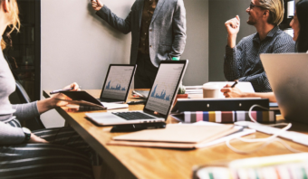 Cuatro personas se sientan en una mesa de conferencias con computadoras portátiles que muestran gráficos mientras una persona está de pie frente a una pizarra, presentando o discutiendo la importancia del Endomarketing en un entorno de oficina moderno.