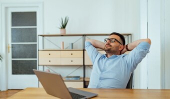 Un hombre disfrutando de sus descansos laborales en su silla con las manos detrás de la cabeza frente a un portátil en una oficina doméstica.