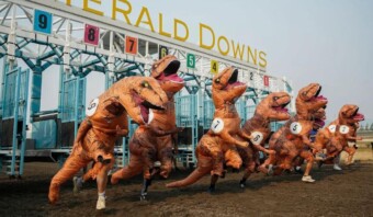 Un grupo de personas disfrazadas de dinosaurios corren frente a una pista de carreras durante las elecciones.