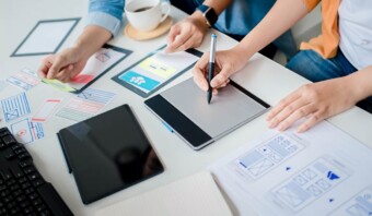 Dos personas colaborando con creatividad en un proyecto de diseño web con tabletas digitales, maquetas de papel y un teclado sobre una mesa blanca.