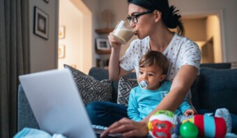 Una mujer bebiendo un vaso de leche mientras trabaja en una computadora portátil, con un niño pequeño en su regazo usando un chupete, en el interior.