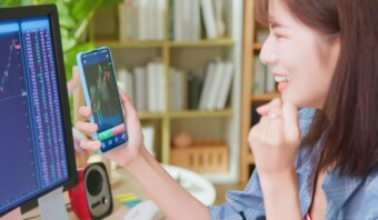 Mujer joven sonriendo mientras mira los gráficos del mercado de valores en su teléfono inteligente, demostrando incrementalidad, con un monitor de computadora mostrando más datos en el fondo.
