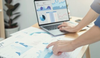 Persona que analiza gráficos de modelado de mezcla de marketing en una computadora portátil y documentos impresos en un escritorio de oficina moderno.