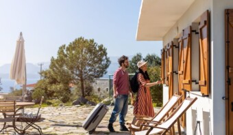 Dos personas, una con maleta y mochila, se acercan a una encantadora casa blanca con contraventanas de madera. El acogedor Airbnb cuenta con una zona de estar al aire libre con sillas de madera y una mesa con sombrilla.