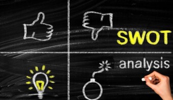 Una pizarra dividida en cuatro secciones con dibujos: un pulgar hacia arriba, un pulgar hacia abajo, "FODA", una bombilla y una bomba. Una mano que sostiene una tiza escribe "análisis" en la sección inferior derecha.