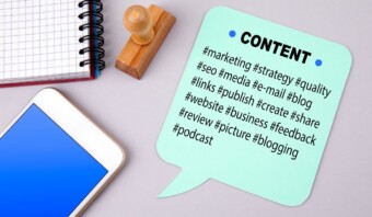 Un bocadillo con la etiqueta "contenido" con hashtags relacionados con el marketing junto a una libreta, un bolígrafo y un sello sobre una superficie gris.