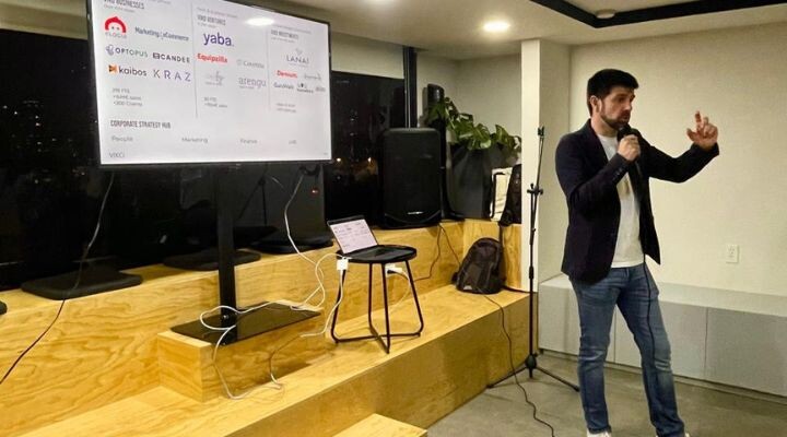 Un hombre se encuentra junto a una pantalla que muestra los logotipos de empresas, incluida Elogia, y habla a través de un micrófono en un espacio de presentación interior moderno.