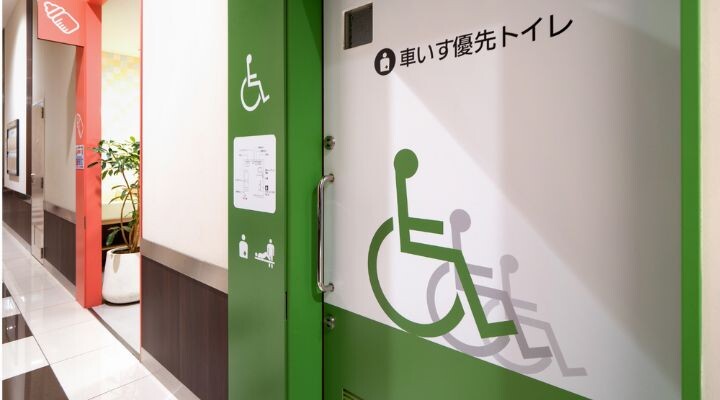 Baño accesible con una puerta verde que presenta un símbolo de silla de ruedas y texto en japonés, ubicado en un pasillo interior limpio, uno de los muchos espacios instagrameables, perfectos para fotos memorables.