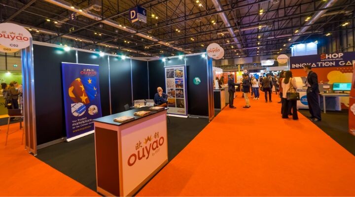 Sala de exposiciones con múltiples stands, incluido un destacado stand de Ouyao en primer plano; la gente camina e interactúa en el fondo, explorando varios espacios instagrameables diseñados para oportunidades fotográficas memorables.
