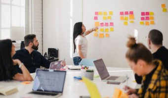 Una mujer, parada frente a una pizarra cubierta de notas adhesivas de colores, hace presentaciones ante cuatro colegas sentados en una sala de reuniones y maneja el estrés del equipo con su enfoque organizado.