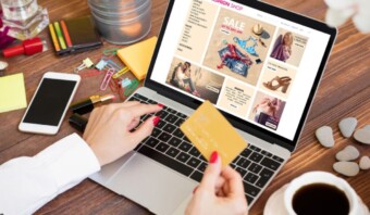 Una persona compra ropa en línea usando una computadora portátil, entusiasmada con los carteles de Venta Caliente que aparecen en la pantalla. Tienen una tarjeta de crédito, listos para realizar compras en diversos artículos de moda. La escena incluye material de oficina y una taza de café, lo que sugiere una sesión de compras espontánea durante el horario laboral.