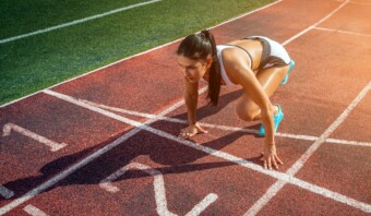 Una mujer vestida con ropa deportiva, que encarna el espíritu del marketing olímpico, está parada en la línea de salida de una pista, lista para correr. Ella está en posición agachada con una rodilla en el suelo y las manos apoyadas en la pista.