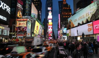 Una concurrida escena callejera en Times Square por la noche, con vallas publicitarias brillantes, numerosos peatones y automóviles, incluidos taxis amarillos, en movimiento.